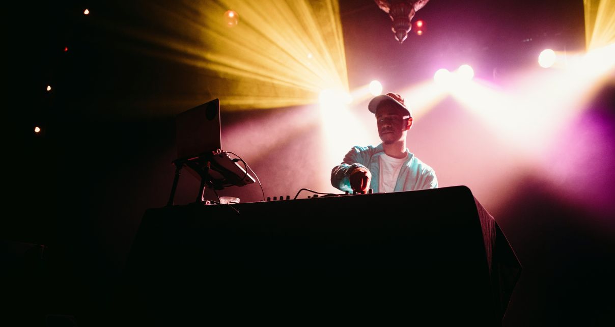 DJ at a club looking at laptop with yellow and pink lighting behind him